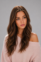 Woman wearing Bradley heat friendly synthetic long wavy wig in color sunlit brunette and a light pink sweater against a light grey background. Front view.