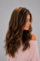 Woman wearing Bradley heat friendly synthetic long wavy wig in color sunlit brunette and a light pink sweater against a light grey background. Right side view.