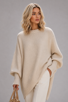 Woman wearing Brielle synthetic wig by PUSH in color golden champagne rooted with a light tan sweater and white pants against a light grey background. 