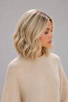 Woman wearing Brielle synthetic wig by PUSH in color golden champagne rooted with a light tan sweater against a light grey background. Right side view.