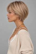Woman wearing Emma Human Hair Wig by PUSH in color Light Champagne Crème Rooted with a cream v-neck sweater against a light grey background-side view