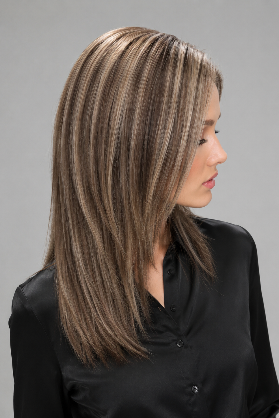Woman wearing Evangeline heat friendly synthetic long wig in color cashmere bronde and a button down black blouse against a light grey background. Right side view.