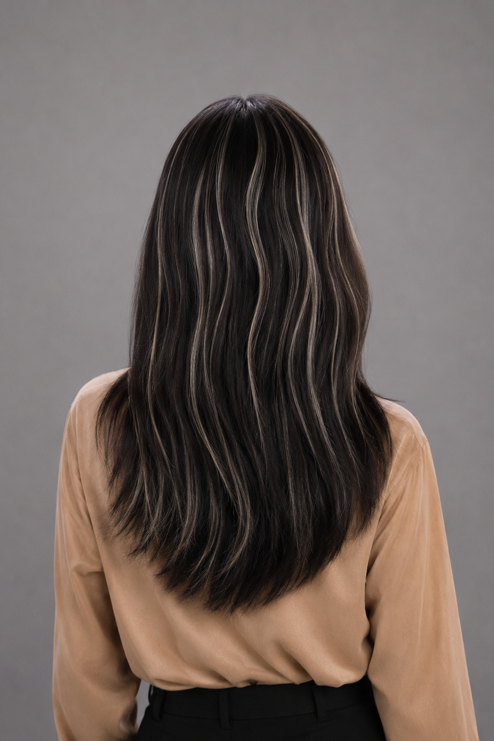 Woman wearing Gisele Human Hair Wig by PUSH in color Étoile Nocturne, a deep and rich off black base with chunky blonde highlights throughout and a tan blouse against a light grey background. Back view.