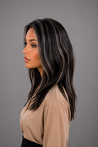 Woman wearing Gisele Human Hair Wig by PUSH in color Étoile Nocturne, a deep and rich off black base with chunky blonde highlights throughout and a tan blouse against a light grey background. Right side view.