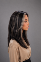 Woman wearing Gisele Human Hair Wig by PUSH in color Étoile Nocturne, a deep and rich off black base with chunky blonde highlights throughout and a tan blouse against a light grey background. Left side view.