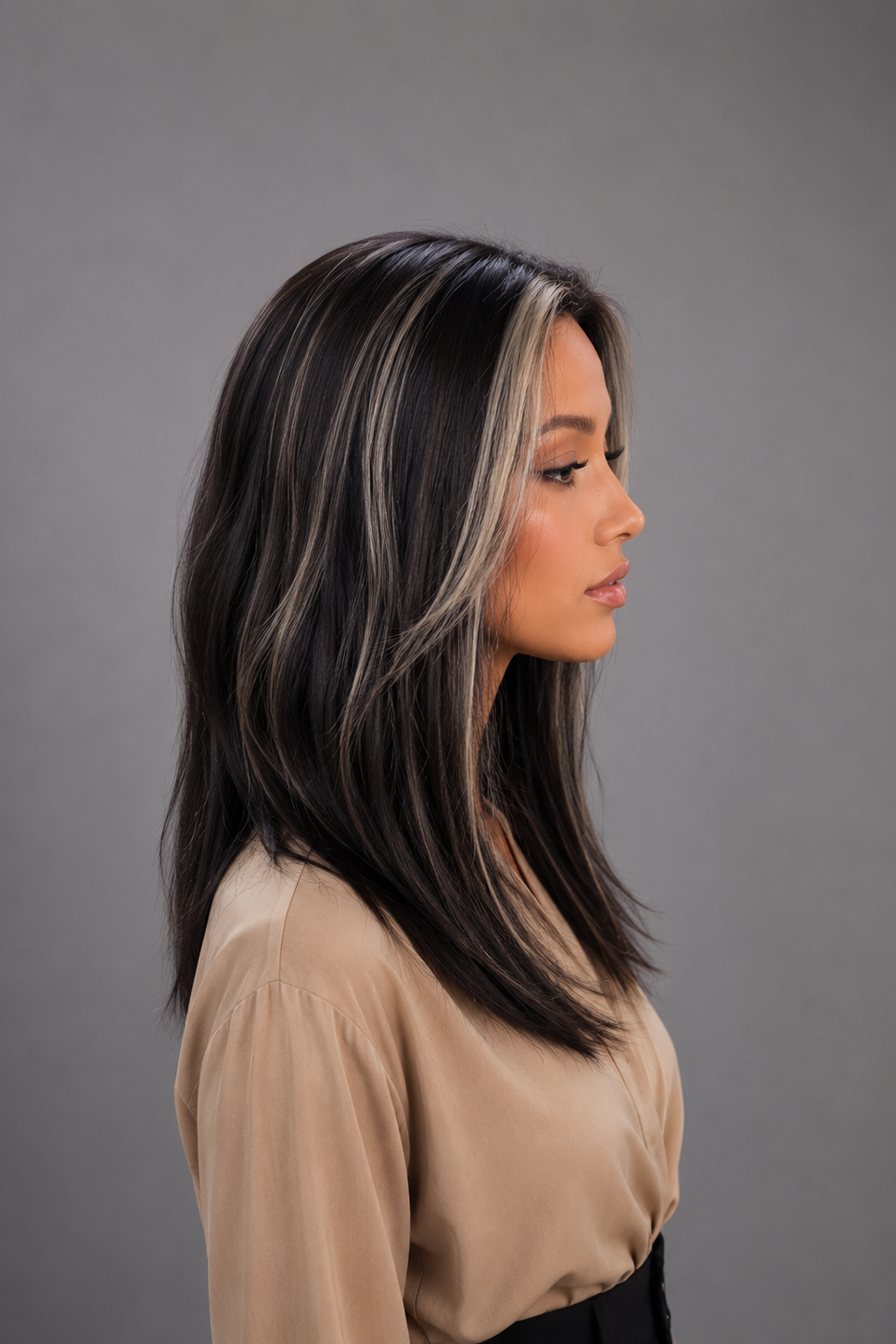 Woman wearing Gisele Human Hair Wig by PUSH in color Étoile Nocturne, a deep and rich off black base with chunky blonde highlights throughout and a tan blouse against a light grey background. Left side view.