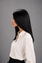 Woman wearing Jacey human hair wig by PUSH in color Noir with cream button down blouse and black pants with a light grey background. Left side view.