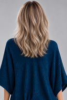 A woman wearing Keeley heat friendly synthetic wavy wig by PUSH Hair in color champagne rooted with a v-neck navy top and a silver necklace on against a light grey background-back view.