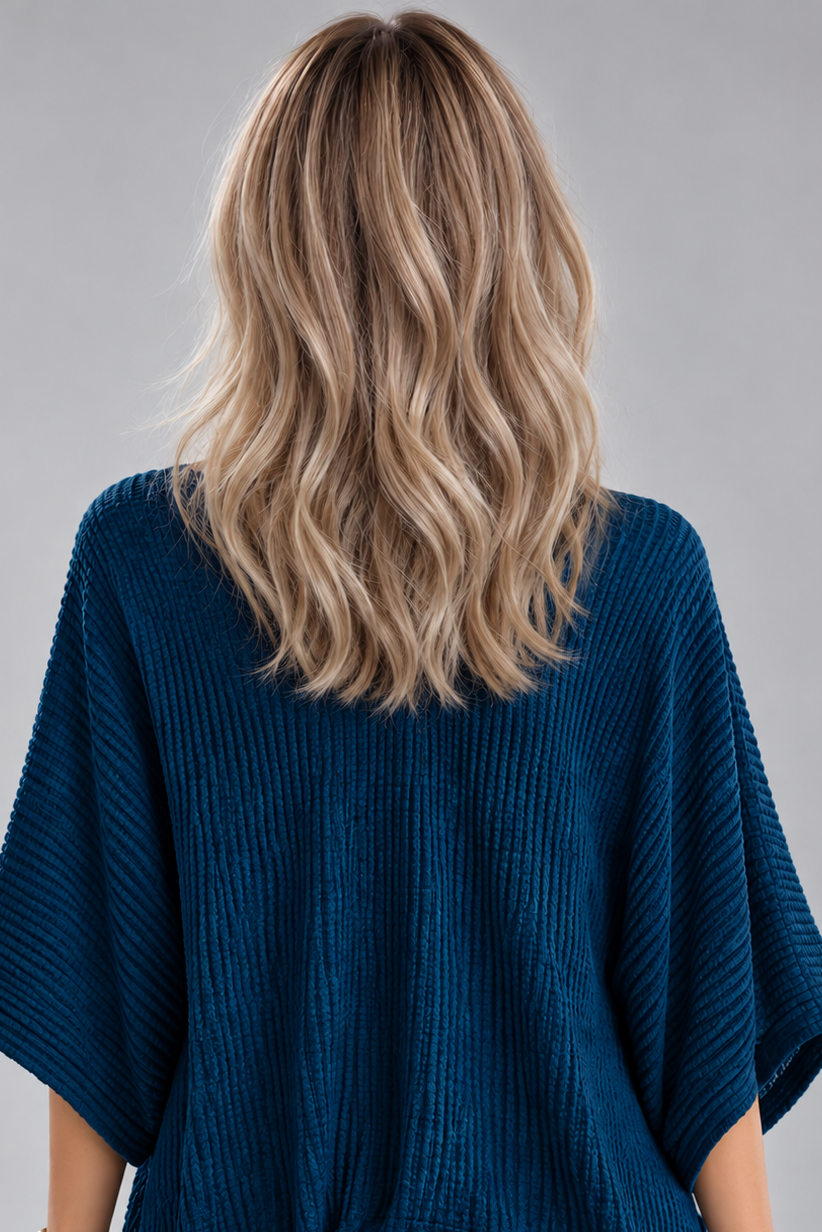 A woman wearing Keeley heat friendly synthetic wavy wig by PUSH Hair in color champagne rooted with a v-neck navy top and a silver necklace on against a light grey background-back view.