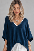 A woman wearing Keeley heat friendly synthetic wavy wig by PUSH Hair in color champagne rooted with a v-neck navy top and a silver necklace on against a light grey background-front view.