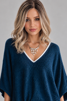 A woman wearing Keeley heat friendly synthetic wavy wig by PUSH Hair in color champagne rooted with a v-neck navy top and a silver necklace on against a light grey background-front view.