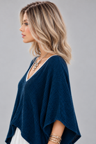 A woman wearing Keeley heat friendly synthetic wavy wig by PUSH Hair in color champagne rooted with a v-neck navy top and a silver necklace on against a light grey background-left side view.