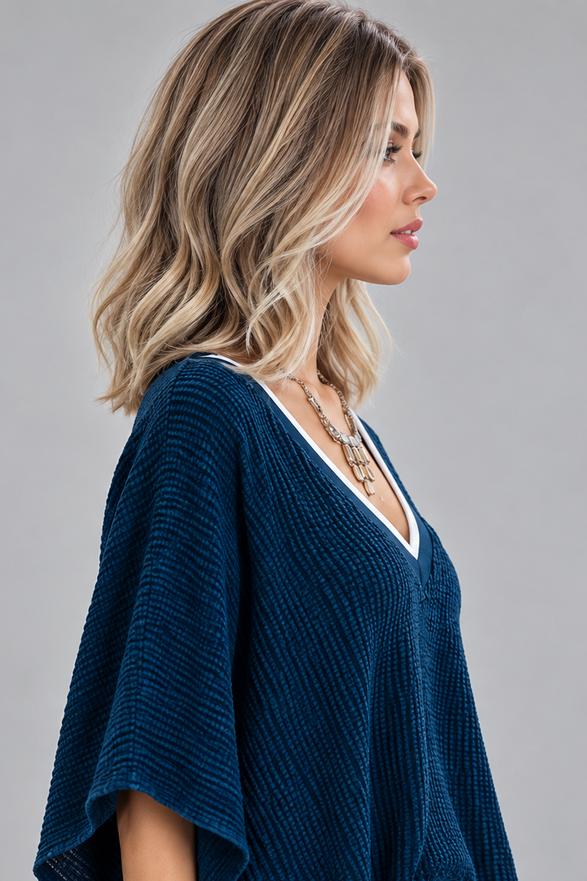 A woman wearing Keeley heat friendly synthetic wavy wig by PUSH Hair in color champagne rooted with a v-neck navy top and a silver necklace on against a light grey background-right side view.