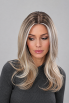 Woman wearing Olivia long, wavy, synthetic wig by PUSH in color pearl silk rooted with a long sleeve charcoal sweater against a light grey background - front view.