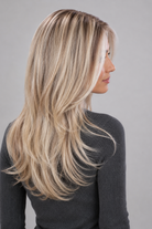 Woman wearing Olivia long, wavy, synthetic wig by PUSH in color pearl silk rooted with a long sleeve charcoal sweater against a light grey background - right side view.