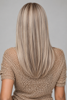 A woman wearing a cross over tan top with necklace and Remmie Human Hair Wig by PUSH Hair against a light grey background - back view
