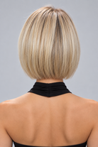 A woman wearing River synthetic wig by PUSH Hair in a blonde rooted shade and a black sleeveless top with gold earrings against a light grey background - back view