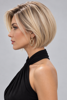A woman wearing River synthetic wig by PUSH Hair in a blonde rooted shade and a black sleeveless top with gold earrings against a light grey background - back view