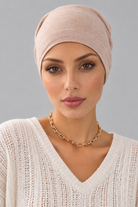 A woman wearing a soft amber melange bamboo head covering or turban-style cap, paired with a cream knit V-neck sweater and a gold chain necklace, against a plain light gray studio background. Front view