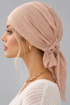 A woman wearing a soft amber melange bamboo head covering or turban-style cap, paired with a cream knit V-neck sweater and a gold chain necklace, against a plain light gray studio background. side view