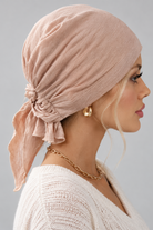 A woman wearing a soft amber melange bamboo head covering or turban-style cap, paired with a cream knit V-neck sweater and a gold chain necklace, against a plain light gray studio background. side view
