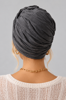 A woman wearing the Wisdom Bamboo Turban in color Anthracite Melange styled with gold hoop earrings and a gold chain necklace against a solid gray background. Back view.