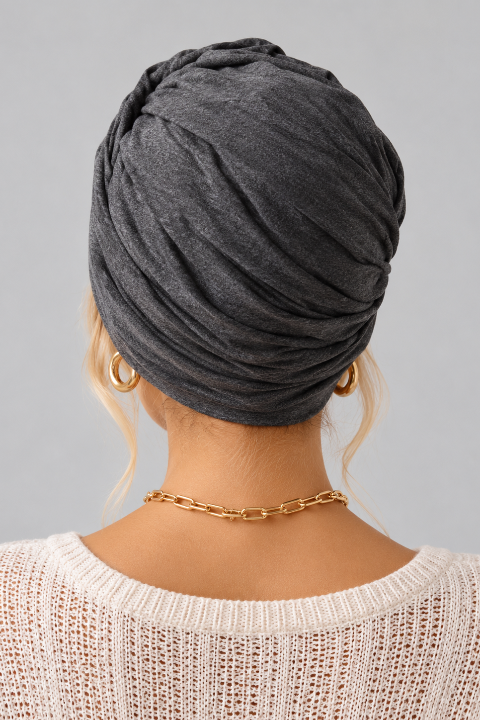 A woman wearing the Wisdom Bamboo Turban in color Anthracite Melange styled with gold hoop earrings and a gold chain necklace against a solid gray background. Back view.