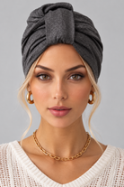 A woman wearing the Wisdom Bamboo Turban in color Anthracite Melange styled with gold hoop earrings and a gold chain necklace against a solid gray background. Front view.