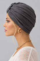 A woman wearing the Wisdom Bamboo Turban in color Anthracite Melange styled with gold hoop earrings and a gold chain necklace against a solid gray background. Left side view.