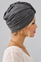 A woman wearing the Wisdom Bamboo Turban in color Anthracite Melange styled with gold hoop earrings and a gold chain necklace against a solid gray background. Right side view.